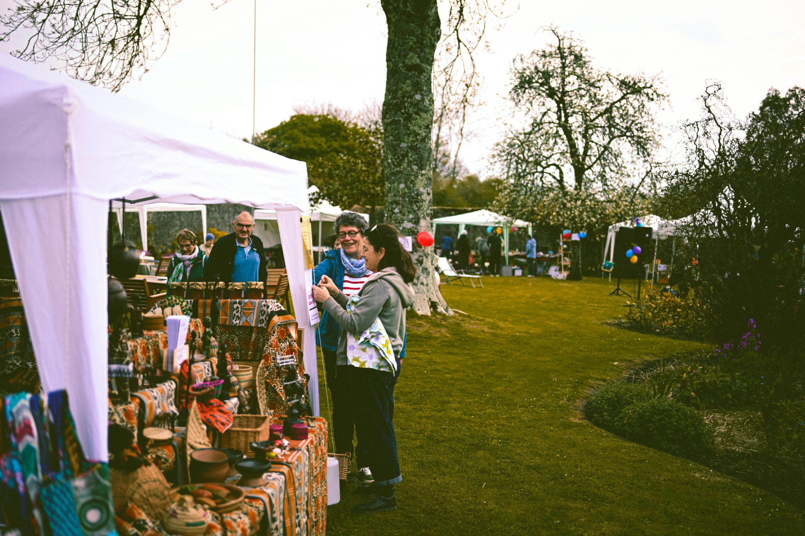 A lively outdoor market with people browsing stalls and tents in a park setting.