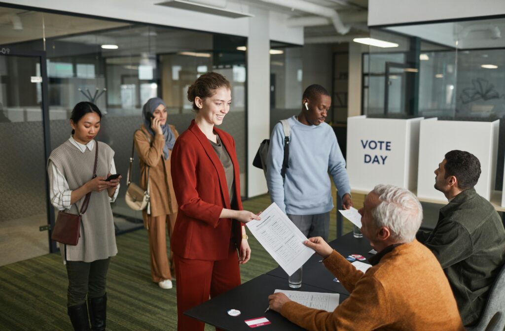 A diverse group of adults participating in an indoor voting process, engaging with election officials.
